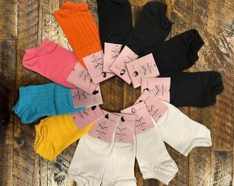 Puede incluir: Una colección de calcetines coloridos en varios tonos, incluyendo rosa, naranja, negro, blanco, amarillo y azul. Cada par está cuidadosamente dispuesto y etiquetado con una etiqueta rosa. Los calcetines se exhiben sobre una superficie de madera.