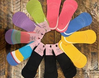May include: A collection of no-show socks in various colors, including pink, blue, yellow, green, purple, and black. The socks are arranged in a circular pattern on a wooden surface. Each pair has a tag attached.