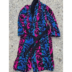 May include: A plush bathrobe featuring a vibrant, swirling pattern in shades of hot pink, bright blue, and teal against a black background. The robe includes a solid black shawl collar, long sleeves, and a matching black tie belt at the waist.