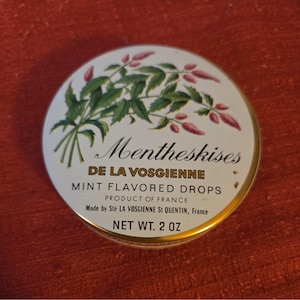 Puede incluir: Una lata metálica redonda de caramelos de menta Mentheskises de la Vosgienne. La tapa blanca presenta una ilustración botánica verde de hojas de menta y flores rosas. El texto incluye: Mentheskises DE LA VOSGIENNE, MINT FLAVORED DROPS, PRODUCT OF FRANCE, Made by Sté LA VOSGIENNE St QUENTIN, France, y NET WT. 2 OZ.