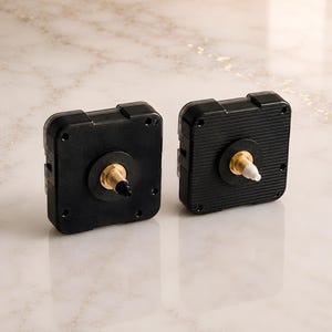 May include: Two black square clock movement mechanisms with brass-colored shafts. The left unit features a black tip on the shaft, while the right unit has a white tip. Both devices have a textured plastic casing with four corner screw holes.