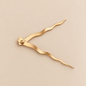 May include: A gold-toned metal hair accessory featuring two long, slender, wavy prongs connected at a single pivot point. The design consists of two curved, serpentine arms that extend outward from a central circular fastener, creating a V-shape.