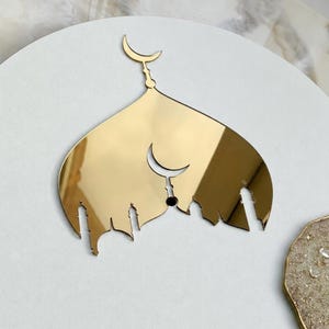 May include: A gold-colored, reflective wall decoration shaped like a mosque silhouette with a pointed dome and minarets. A crescent moon symbol is positioned at the top peak and another smaller crescent moon is centered within the main structure.