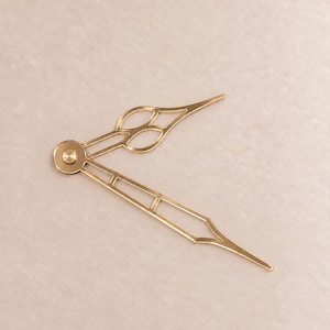 May include: A pair of gold-colored metal clock hands featuring an ornate, open-work design. The hour hand has a decorative, diamond-shaped cutout near the center, while the minute hand features a long, slender, open-frame structure.