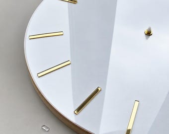May include: A circular wall clock featuring a smooth white face with reflective gold-toned rectangular hour markers. The clock has a central gold-toned mounting post for hands and a light-colored wooden frame edge visible along the curved perimeter.