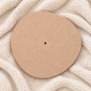 May include: A circular, light brown wooden disc with a smooth, flat surface and a single, small, centered hole. The object is made of a compressed wood material, featuring a uniform, neutral tan color throughout its entire circular shape.