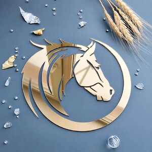 May include: A gold-colored, reflective wall decoration shaped like a horse head silhouette enclosed within a circular frame. The design features stylized, flowing lines representing the mane and neck, creating a polished, mirror-like surface finish.
