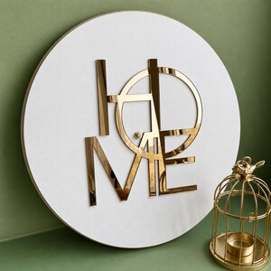 May include: A circular white wall plaque featuring the word HOME in reflective gold-toned lettering. The letters are arranged in a stylized, geometric layout where the O and M share structural lines. A small gold birdcage sits to the right.