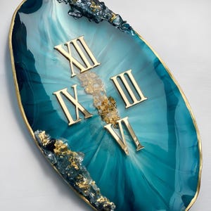 May include: An oval wall clock featuring a teal and blue resin surface with a swirling pattern. The clock face displays gold Roman numerals XII, III, VI, and IX. The edges are accented with gold leaf and dark blue crystalline resin textures.