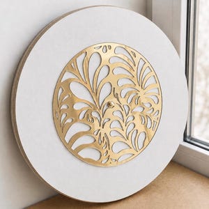 May include: A circular wall clock featuring a white face and a central gold-colored metallic overlay with an intricate, organic cutout pattern. The clock has a smooth, light-colored wooden edge and a small gold-colored center pin for the hands.