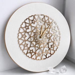 May include: A circular wall clock featuring a white face and a central gold-colored metal overlay composed of an intricate, repeating heart-shaped pattern. The clock includes two slender gold-colored hands at the center, set against a clean, flat background.