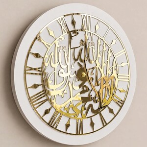 May include: A circular wall clock with a white frame and a gold-colored metallic face. The clock features intricate Arabic calligraphy in the center and gold Roman numerals around the perimeter, with two thin gold hands pointing toward the center.