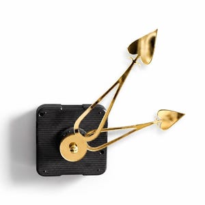 May include: A black square clock movement mechanism featuring two metallic gold clock hands. The hands are shaped like elongated hearts at the tips, extending from a central circular gold cap attached to the black plastic housing unit.