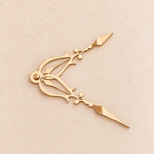 May include: A pair of gold-colored metal clock hands featuring an ornate, openwork design with pointed tips and circular accents. The hands are connected at a central circular hub, forming an angled shape with intricate, symmetrical cutouts.