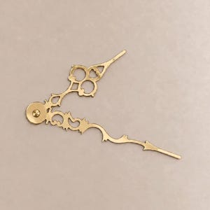 May include: A pair of gold-colored metal clock hands featuring an ornate, decorative cutout design. The hands are connected at a central circular hub, with one hand shorter than the other, both displaying intricate, scrolling filigree patterns.