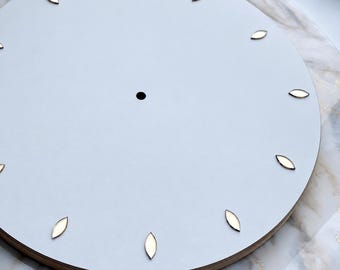 May include: A circular light gray clock face featuring a central hole for a mechanism and twelve small, metallic, leaf-shaped hour markers arranged around the perimeter. The clock face has a smooth, flat surface and a visible wooden edge.