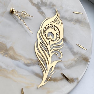 May include: A wall clock featuring a circular face with a white and gold marble pattern. The clock includes gold-colored metal hands and a large, ornate, gold-colored metal peacock feather cutout that serves as a decorative central element.