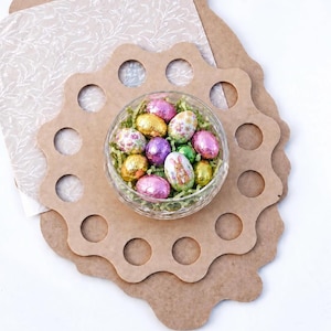 May include: A clear glass bowl filled with colorful foil-wrapped chocolate eggs, including one with a rabbit illustration, resting on a light brown wooden display board featuring a scalloped edge and twelve circular cutouts for holding eggs.