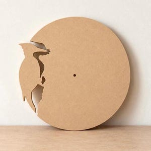 May include: A circular light brown wooden clock face blank featuring a laser-cut silhouette of a woodpecker on the left side. The flat, unfinished wood surface has a small, centered hole for a clock mechanism, showcasing a smooth texture.