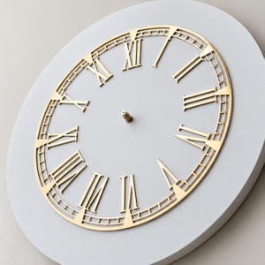 May include: A circular wall clock face featuring a smooth light gray base and a metallic gold overlay. The overlay displays large Roman numerals and a thin outer ring with grid-like detailing. A small metal pin is centered on the clock face.