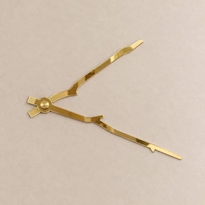 May include: A pair of gold-colored clock hands with a branch-like, organic shape. The two hands are connected at a central circular hub, featuring irregular, textured edges that mimic the appearance of small tree twigs or wooden branches.