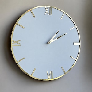 May include: A round wall clock featuring a matte white face and a polished gold-tone metal frame. The clock displays gold-tone Roman numerals at the twelve, three, six, and nine o'clock positions, with simple gold-tone hour and minute hands.