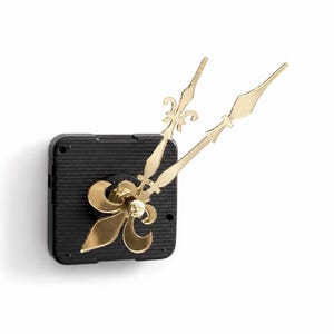 May include: A black square clock movement mechanism featuring two ornate gold-colored clock hands. The hands are designed with a fleur-de-lis motif at the center and decorative pointed tips, providing a metallic finish to the clock assembly.