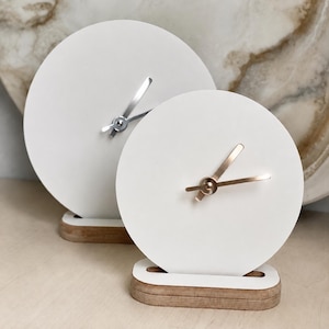 May include: Two circular table clocks with smooth white faces and wooden bases. The larger clock features silver-toned hands, while the smaller clock in the foreground has rose gold-toned hands. Both clocks are mounted on rectangular wooden stands.