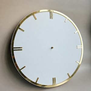 May include: A circular wall clock featuring a plain white face and a polished gold-tone metal rim. The clock face includes thin gold-tone hour markers, with double markers at the twelve and six o'clock positions, and a small gold centre pin.