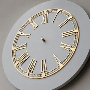 May include: A circular wall clock face featuring a smooth light grey background and a gold-coloured metal overlay. The overlay displays large Roman numerals and a thin outer ring with tick marks. A small metal pin is centred on the clock face.