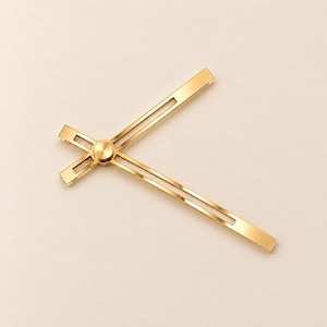May include: A set of two gold-toned metal clock hands featuring a slender, open-frame design with a central circular mounting cap. The hands are positioned at an angle, showcasing a polished metallic finish and a clean, linear structure.