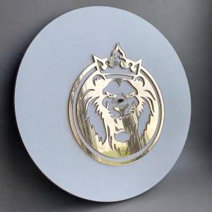 May include: A circular wall clock featuring a matte light gray face and a centered, reflective gold-toned cutout of a lion head wearing a crown. The lion design is enclosed within a thin gold circular frame, creating a layered, metallic aesthetic.