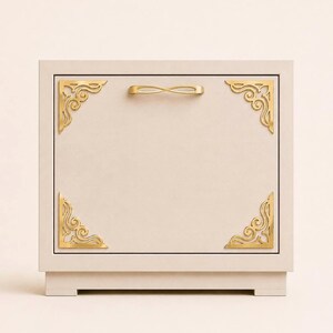 May include: A square, light beige storage box featuring a central horizontal gold handle and four decorative gold-toned metal corner brackets with intricate scrollwork patterns attached to the front panel of the box.