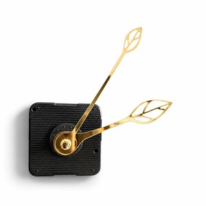 May include: A black square clock mechanism featuring two gold-colored clock hands shaped like open-work leaves. The central hub is circular and metallic gold, connecting the two leaf-patterned hands to the textured black plastic housing.