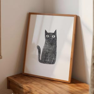 Könnte beinhalten: Ein gerahmter Kunstdruck mit einer Illustration einer schwarzen Katze. Die Katze ist mit strukturierten Linien gezeichnet und hat große, weite Augen. Das Kunstwerk ist vor einem weißen Hintergrund platziert und in einem hellbraunen Holzrahmen gerahmt. Der Druck wird auf einer Holzoberfläche präsentiert.