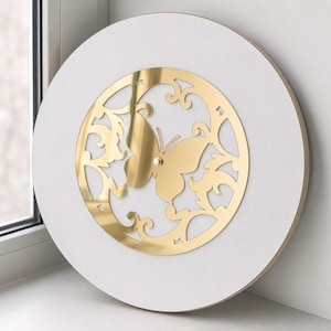 May include: A circular wall clock with a matte white face and a central gold-toned metallic inlay. The inlay features a cutout design of a butterfly surrounded by swirling filigree patterns. A small silver-colored center post holds the clock hands.