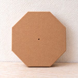 May include: A light brown octagonal wooden board with a smooth, flat surface and a single small circular hole centered in the middle. The object features clean, straight edges and a uniform, solid tan color throughout its entire structure.