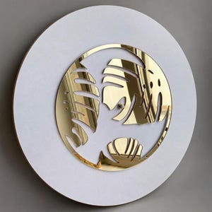 May include: A circular wall art piece featuring a white matte base with a centred, reflective gold-toned circular inlay. The gold inlay is intricately cut to display stylised monstera leaf silhouettes, creating a high-contrast, layered appearance.