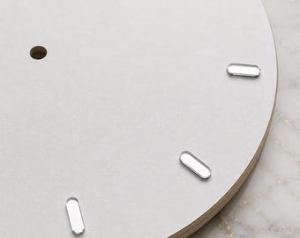 May include: A circular clock face featuring a smooth, light gray surface with a central hole for a mechanism. Four small, metallic, pill-shaped markers are positioned along the edge to indicate time, set against a light-colored wooden base.
