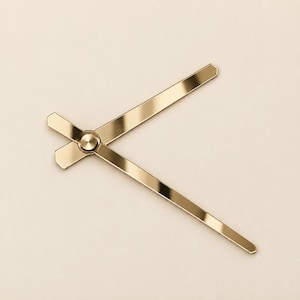 May include: A pair of metallic gold clock hands featuring a polished finish and a central circular mounting cap. The hands are straight with pointed ends, designed for a clock mechanism, and are positioned at an angle to each other.