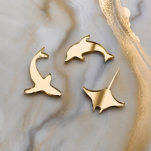 May include: Three gold-colored mirror-finish acrylic cutouts shaped like marine animals. The set includes a dolphin, a shark, and a manta ray, each featuring a smooth, reflective surface that captures light and creates a distinct metallic appearance.