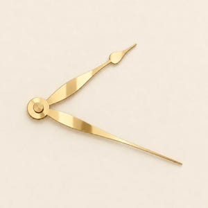 May include: A pair of gold-colored clock hands featuring a central circular mounting hub. The shorter hand has a tapered, leaf-like shape with a pointed tip, while the longer hand is slender and straight, extending to a fine point.