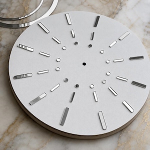 May include: A circular white clock face featuring a central hole, an inner ring of small silver circular markers, and an outer ring of elongated silver rectangular markers. A separate silver circular ring is positioned near the top edge.