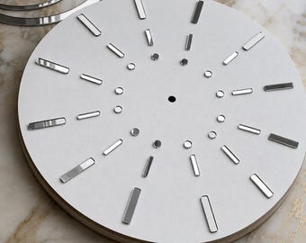 May include: A circular white clock face featuring a central hole, twelve rectangular silver hour markers arranged in a radial pattern, and an inner ring of twelve small circular silver markers. A separate silver ring sits partially visible above.