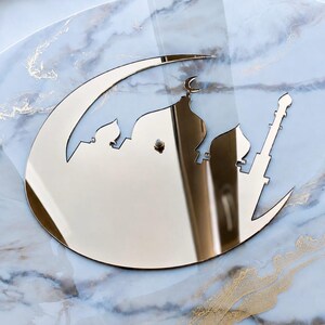 May include: A gold-colored reflective wall clock featuring a crescent moon shape with a silhouette of a mosque, including multiple domes and a tall minaret. The clock face has a central mechanism post and a smooth, polished metallic surface finish.