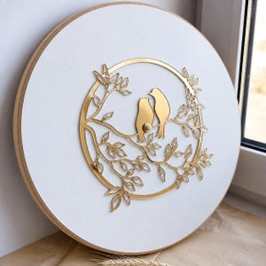 May include: A circular wall clock with a white face and a light wood frame. Centered on the face is a gold-colored metal cutout featuring two birds perched on a branch within a circular border, surrounded by intricate leaf and vine details.