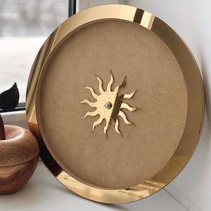 May include: A circular wall clock featuring a polished gold-tone metal frame and a plain, light brown fiberboard face. At the center, a gold-tone sun-shaped emblem with wavy rays serves as the focal point, lacking visible clock hands or numerals.