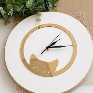 May include: A round white wall clock featuring a gold-colored mirrored circular frame and a stylized cat head silhouette at the bottom. The clock has three black hands positioned in the center, with the cat's eyes cut out from the gold surface.
