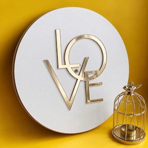 May include: A circular white wall plaque featuring the word LOVE in large, reflective gold-toned metallic letters. The letters are arranged in a stacked, stylized format. Beside the plaque sits a small, gold-toned metal birdcage candle holder.
