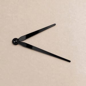 May include: A pair of black clock hands featuring a smooth, tapered design with a central circular mounting hub. The two hands are positioned at an angle, extending outward from the center point, showcasing a sleek, solid black finish.